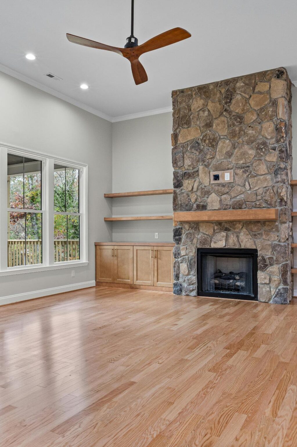 Empty living room with light wood floors, stone fireplace, built-in wooden shelves, large windows, and ceiling fan in a neutral modern design.