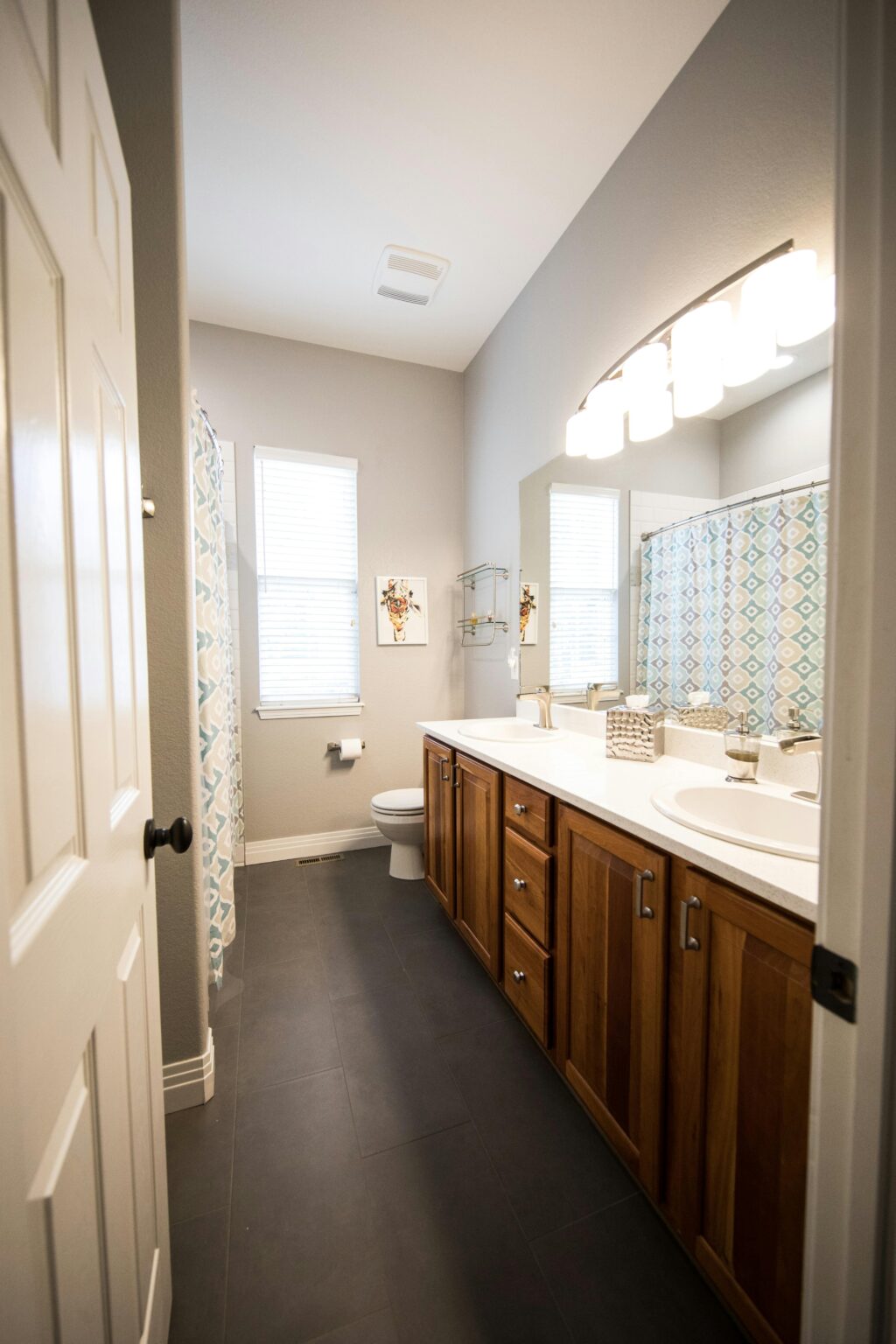 Long narrow bathroom with double vanity featuring wooden cabinets, large wall mirror, and overhead lighting; toilet and window at the far end with light gray walls and dark flooring. The best house cleaning service in Exeter.